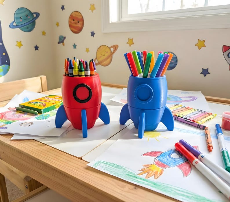 Red and blue 3D-printed rocket crayon holders filled with art supplies on a children’s drawing table